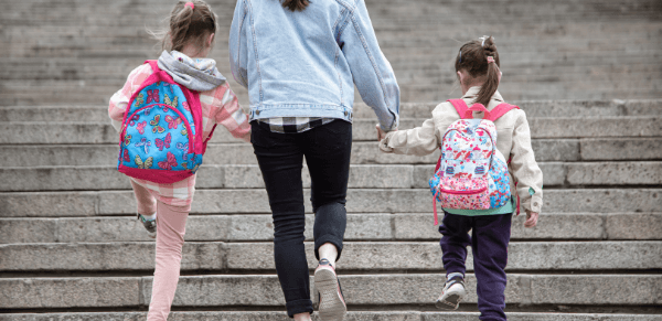 Comunicação entre família e escola: imagem de mãe segurando na mão de suas duas filhas que estão com mochilas nas costas.