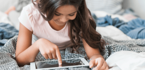Vantagens e desvantagens do ensino híbrido: imagem de uma menina estudando com seu tablet na cama do seu quarto